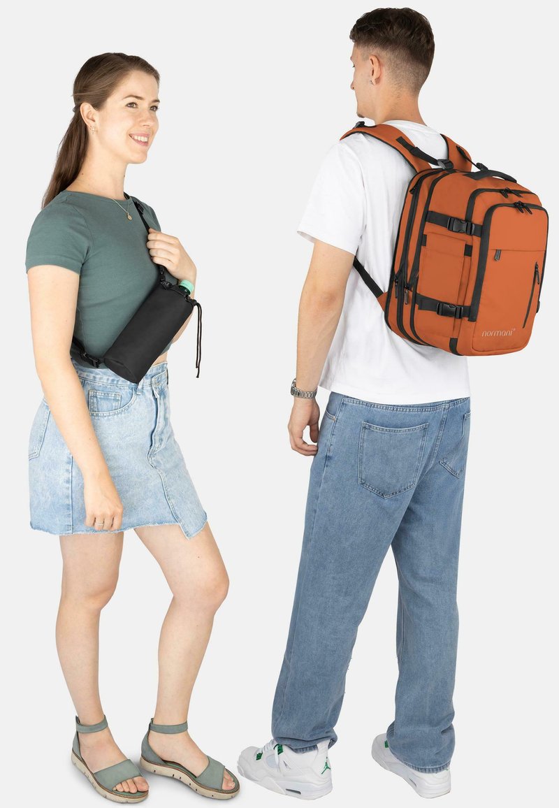 Schwarze Umhängetasche mit Kordelzug, getragen von einer Frau in grünem Oberteil und Jeansrock; Mann in weißem T-Shirt und orangefarbenem Rucksack, blaue Jeans.