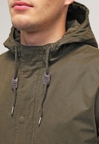 Olivgrön huva jacka med en texturerad yta, med dragkedja och justerbara snoddar, samt logotypmedaljonger i metall.