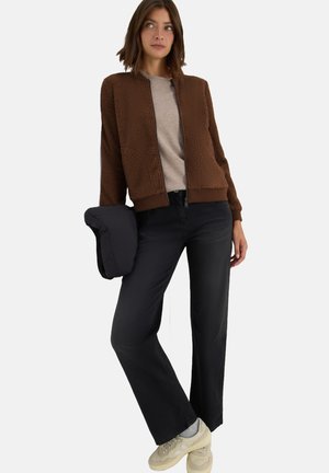 Veste bombardier marron en peluche, chemise grise, jean noir à jambes larges et baskets beiges. La veste présente un ourlet côtelé et une fermeture éclair.
