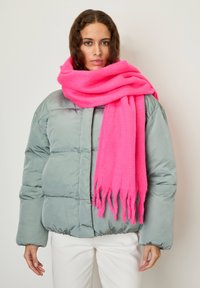 Woman with wavy brown hair wearing a gray puffer jacket, white pants, and a large bright pink fringed scarf wrapped around her neck.