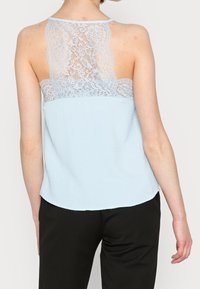 Ljusblå linne med spetsdetaljer bak, slät tyg, rundad fåll och spaghetti-remmar; elegant spetsdekoration över övre ryggen.