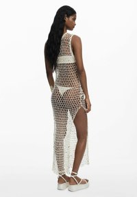Dlhé krémové háčkované šaty s geometrickým vzorom, s vysokým rozparkem a odhaleným chrbtom. V kombinácii s bielymi platformovými sandálmi.