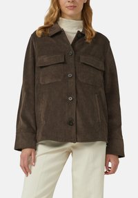 Vrouw in een bruine corduroy button-up jas met klepzakken op de borst, over een lichte coltrui en crèmekleurige broek.