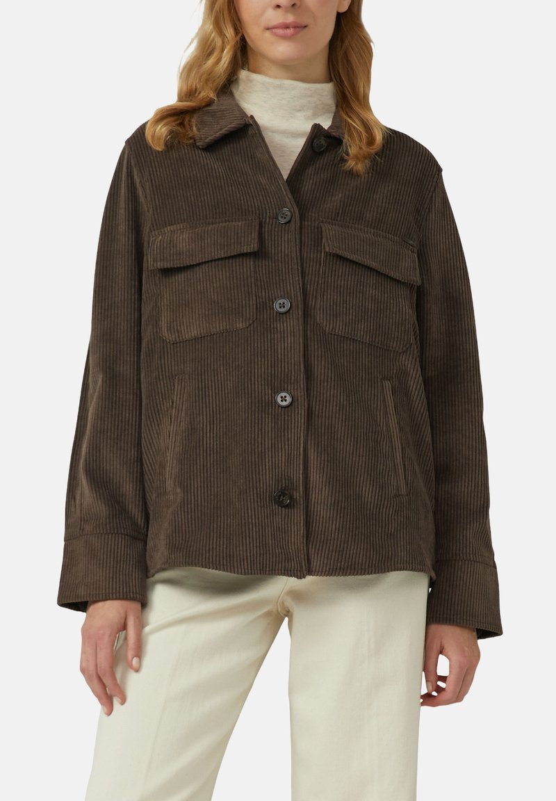 Vrouw in een bruine corduroy button-up jas met klepzakken op de borst, over een lichte coltrui en crèmekleurige broek.