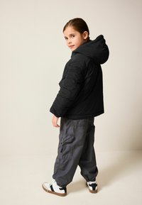 Schwarze gepolsterte Jacke mit Kapuze, getragen mit grauen Cargo-Hosen mit Taschen und weißen Sneakern mit schwarzen Akzenten. Einfaches und funktionales Design.