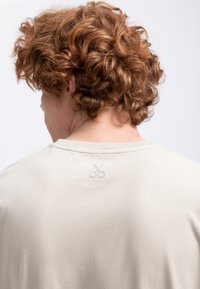 Lichtbeige t-shirt met een ronde hals, korte mouwen en een klein geborduurd schaar detail aan de achterkant vlakbij de kraag.