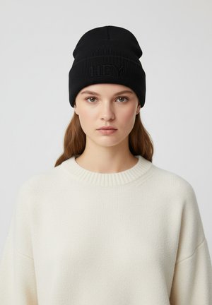 Bonnet tricoté noir avec une texture côtelée et un logo "HBY" en relief. Porté avec un pull beige clair à col rond et coupe loose.
