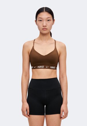 Un mannequin se tient devant un fond clair, vêtu d’une brassière de sport Nike marron et d’un short noir ajusté.