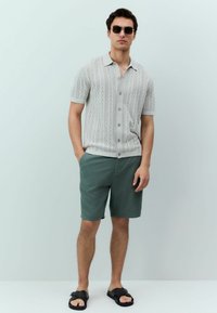 Cămașă scurtă gri deschis, cu un model texturat, asortată cu pantaloni scurți verzi și sandale negre, purtată de un bărbat care stă în fața unui fundal simplu.