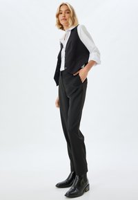 Gilet noir sur une chemise blanche, associé à un pantalon sombre. Textures lisses et coupe ajustée, complétées par des bottines noires avec un accent doré.