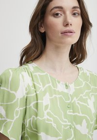 Bluză verde cu un model relaxat, având forme abstracte albe, decolteu rotund și nasturi mici verzi pe față.