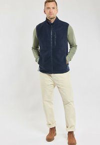 Gilet en polaire bleu marine avec un col haut et une poche zippée, porté par-dessus une chemise à manches longues vert clair et associé à un pantalon beige et des bottes marron.