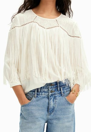 Femme portant un chemisier blanc à franges avec un empiècement brodé et un jean taille haute bleu clair, mains dans les poches, portant des bracelets en or au poignet droit.