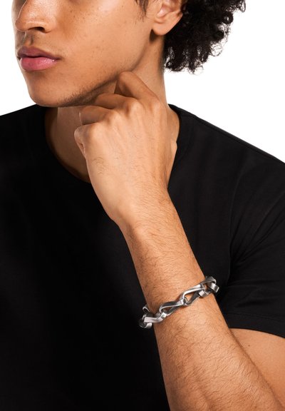Bracelet chaîne en métal argenté avec maillons entrelacés, porté au poignet, assorti à un T-shirt noir. Texture lisse et finition brillante.