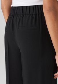 Pantalon noir avec une texture lisse, une taille élastique et une seule poche arrière située sur le côté droit.