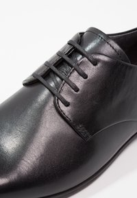 Chaussure en cuir noir avec une finition lisse, un design à lacets et un bout arrondi. Les détails notables incluent des accents cousus et une texture brillante.