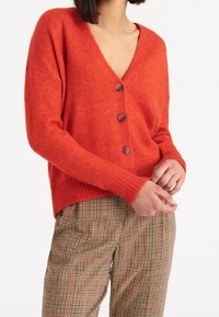Oranžový pletený cardigan s V-výstřihem, zapínáním na knoflíky a žebrovanými manžetami. Nosený přes kostkované hnědé a béžové kalhoty.