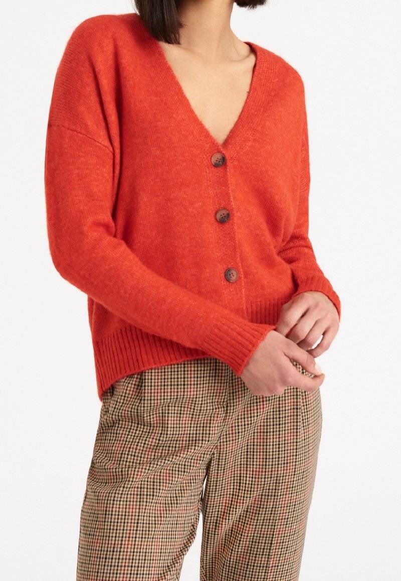 Oranžový pletený cardigan s V-výstřihem, zapínáním na knoflíky a žebrovanými manžetami. Nosený přes kostkované hnědé a béžové kalhoty.