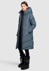 Langer, voluminöser, blau-grauer Mantel mit großer Kapuze, diagonaler Steppung, Frontreißverschluss und seitlichen Taschen. Kombiniert mit schwarzen, isolierten Stiefeln.