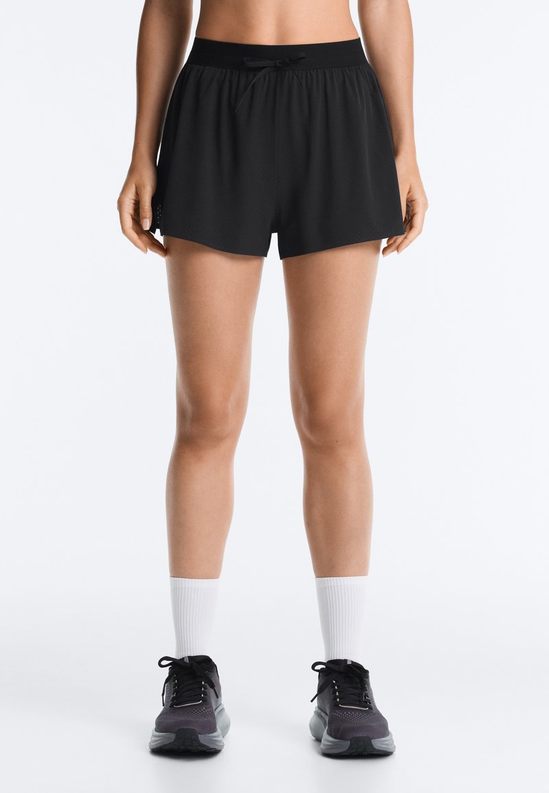 Personne portant un short de sport noir, des chaussettes blanches montantes et des chaussures de course noires, debout devant un fond clair uni.