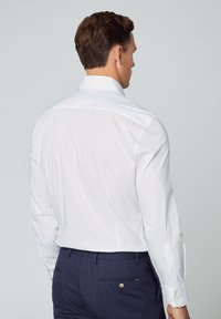 Weißes Langarmhemd mit geradem Schnitt, glatter Textur und Knopfmanschetten, kombiniert mit dunkelkarierten Hose mit einer Gesäßtasche.