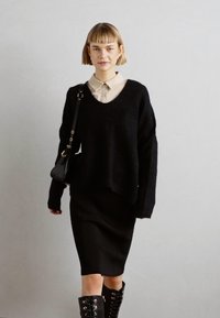 Svart oversized strikkegenser med V-hals, beige skjorte under, svart blyantskjørt og knelang svart snørestøvler.