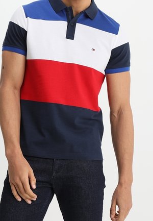 Homme portant un polo à manches courtes avec des rayures horizontales bleues, blanches, rouges et bleu marine et un jean foncé.