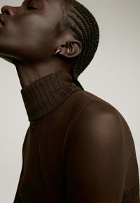 Suéter marrón de cuello alto con un diseño ajustado. La textura suave destaca el escote, complementado por el cabello trenzado a un lado y un sutil arete dorado.