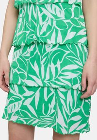 Grüner und weißer gestufter Rock mit Blättermustern, der einen welligen Saum und einen weichen, strukturierten Stoff aufweist. Hände sind sichtbar, die den Rock halten.