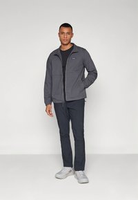 Jachetă gri cu fermoar și guler ridicat, două buzunare frontale, asortată cu pantaloni închiși la culoare și adidași gri, pe un fundal simplu.