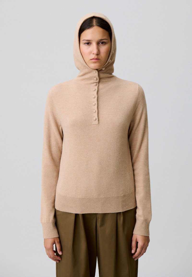 Sweat à capuche tricoté beige avec un col haut et des détails de boutons. Manches et ourlet côtelés, avec une texture douce et une silhouette ajustée.