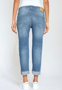 Mujer vestida con jeans azules remangados y zapatillas blancas Reebok, de pie con las manos relajadas a los lados contra un fondo blanco.