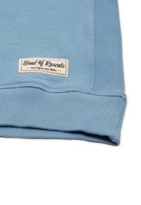 Vaaleansininen ribattu kangas, jossa on valkoinen etiketti, jossa lukee "Band of Rascals Finest Organic Kids Clothes", ommeltu lähelle helmaa.