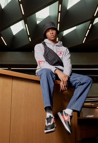 Nike sneakers with black, grey, and red accents; grey hoodie with red logo; black bucket hat; blue jeans; black shoulder bag.
