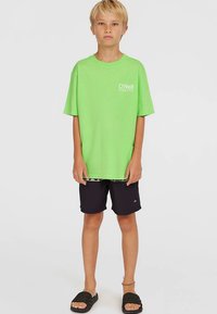 Neonvihreä puuvillainen t-paita valkoisella logolla, mustat uimashortsit ja mustat liukusandaalit; rentoa rantavaatetusta nuorille.