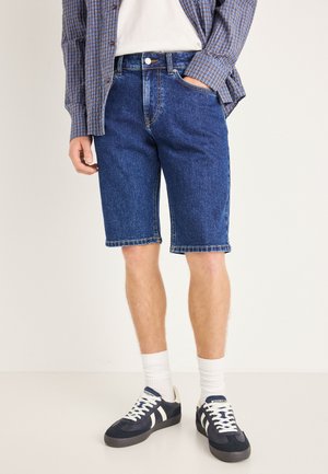 Shorts en denim bleu foncé, avec une coupe droite, des coutures visibles à l'ourlet et deux poches avant, assortis à des baskets noires et des chaussettes blanches.