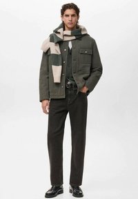 Giovane uomo che indossa una giacca verde, pantaloni neri, scarpe nere e una sciarpa a righe verde e beige drappeggiata sulle spalle.