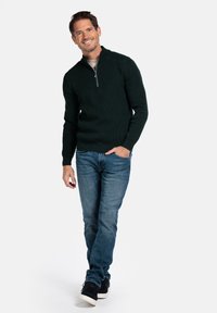 Donkergroene gebreide trui met een halve rits kraag, gedragen over een lichtgekleurde shirt, gecombineerd met blauwe spijkerbroeken en zwarte sneakers.