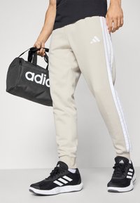 Bežiniai sportiniai kelnių su baltais šoninėmis juostelėmis, juodi sportiniai bateliai su baltais akcentais ir juodas sportinis krepšys su dideliais baltais užrašais.