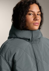 Prošivena jakna u prigušenoj nijansi plave boje, s visokim ovratnikom, dijagonalnim šavovima i glatkom teksturom. Prikaz tkanine i detalja izbliza.