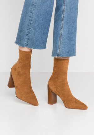 Brune ruskind ankelstøvler med spids tå og blokhæl, båret med cropped blå jeans på et hvidt gulv.