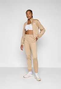 Beige träningsjacka och byxor med vitt logotyp, tillsammans med en vit sport-bh och ljusa sneakers. Slät tyg, figurnära design.