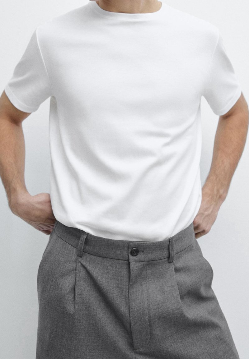 Homme portant un t-shirt blanc uni à manches courtes rentré dans un pantalon gris taille haute, les mains posées sur les hanches.