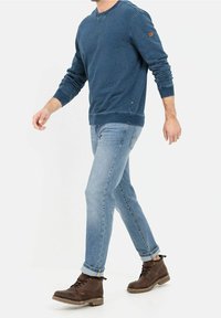 Felpa di cotone blu con scollatura a girocollo, abbinata a jeans azzurri e stivali di pelle marrone. Presenta un piccolo logo sulla manica.