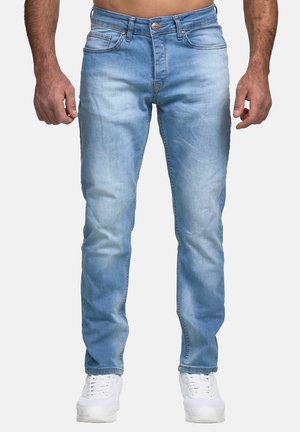 Man die lichtblauwe slim-fit jeans en witte sneakers draagt, staand tegen een effen achtergrond met handen ontspannen langs het lichaam.