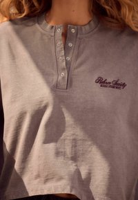 Henley stiilis lühike pluus vaigistatud lavendlivärvuses, ümmarguse kaeluse, viie n�ööbiga loodud ja tume burgundia värviga brodeeritud logoga.
