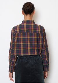 Camicia a maniche lunghe a quadri con motivi neri, rossi, gialli e viola. Polsini con bottoni e dettaglio sulla schiena. Indossata con una gonna in velluto a coste scura.