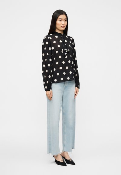 Μαύρο πουκάμισο με πουά σχέδιο και βολάν στο ντεκολτέ, σε συνδυασμό με ανοιχτό μπλε τζιν παντελόνι με φαρδιά μπατζάκια και μαύρες γόβες με μυτερή μύτη. Ομαλή υφή υφάσματος.