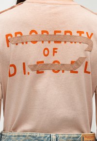 Μπεζ πουκάμισο με πορτοκαλί κείμενο «PROPERTY OF DIESEL», μερικώς καλυμμένο από δύο υφασμάτινες μπεζ λωρίδες, φορεμένο με ανοιχτόχρωμα τζιν παντελόνια.