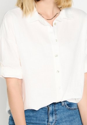 Biała koszula zapinana na guziki z podwiniętymi rękawami, zestawiona z niebieskimi jeansami oraz cienkim srebrnym naszyjnikiem na osobie.